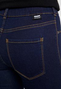 Pantalones vaqueros de mezclilla azul oscuro con costuras amarillas, que cuentan con un bolsillo trasero y una pequeña etiqueta de marca que dice "DRDENIM". Textura suave, diseño ajustado.