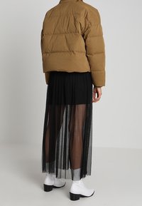 Personne portant une doudoune brune, une longue jupe noire transparente par-dessus un short noir, et des bottines blanches, debout devant un fond uni.