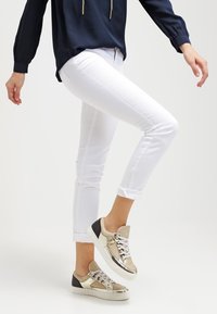 Vita tajta byxor med uppvikta fållar, tillsammans med metalliska guld- och svarta sneakers med snörning och accentdetaljer.