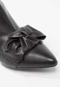 Zapato de cuero negro con punta afilada que presenta un gran lazo decorativo en la parte frontal y costuras visibles a lo largo del costado.