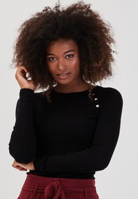 Haut à manches longues noir côtelé avec trois accents de boutons dorés sur l'épaule, présentant une silhouette ajustée et une texture douce.