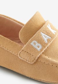 Kohvitahk nahast loafers pehmea tekstuuriga, millel on lai rihm valgete tikanditega sõnadega "BAK." Ümar varvas ja õmmeldud detailid.