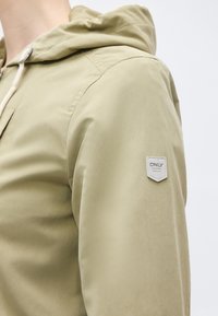 Κοντινό πλάνο ατόμου που φοράει μπεζ μπουφάν με κουκούλα, με κουμπί και λευκή ετικέτα με την επιγραφή "ONLY" στον άνω βραχίονα.