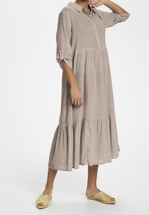 Kvinde iført en løs beige midi-kjole med lagdelt kant, opkørte ærmer og vævede slidser-sandaler, stående mod en ensfarvet hvid baggrund.
