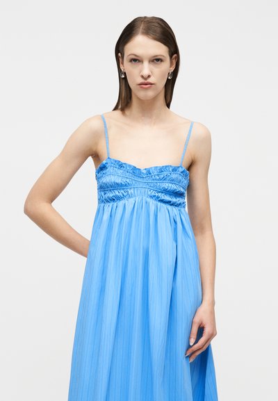 Γυναίκα με φόρεμα χωρίς μανίκια, λαμπερό μπλε, με συγκεντρωμένο μπούστο και λεπτές τιράντες, που στέκεται μπροστά σε απλό ανοιχτό φόντο.