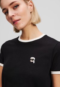 Black t-shirt with a round neck, white trim. Features an embroidered design of a person wearing sunglasses on the chest. Smooth cotton texture.