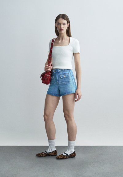 Νεαρή γυναίκα που φοράει λευκό τοπ με κοντά μανίκια, τζιν σορτς με ψηλή μέση, λευκές κάλτσες, λεοπάρ πλατφόρμες και κόκκινη τσάντα ώμου, στέκεται μπροστά σε απλό φόντο.