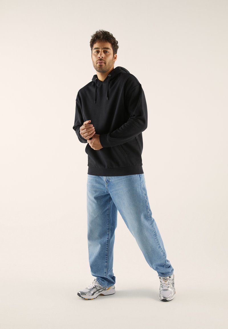 Schwarzer Kapuzenpullover mit Kängurutasche, kombiniert mit hellblauen Jeans und grauen Turnschuhen, vor einem schlichten Hintergrund.