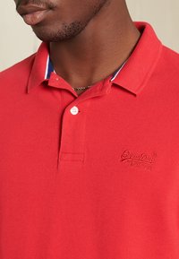 T-shirt polo rouge en tissu texturé, avec patte de boutonnage à deux boutons et logo brodé. Le col présente des accents bleus et blancs.