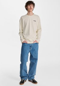 Cremefarbene Langarm-T-Shirts mit schwarzem Logo; locker sitzende blaue Jeans; schwarze Sneakers mit weißen und braunen Akzenten.