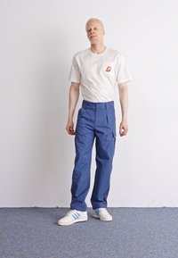 Witte T-shirt met korte mouwen, voorzien van een zak en logo, gecombineerd met blauwe cargo-pantalons. Het model draagt witte sneakers met blauwe strepen. Effen achtergrond.