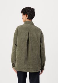 Camisa de sarja verde, corte relaxado, colarinho largo, punhos com botão, textura riscada vertical, detalhe na pala das costas, ligeiramente mais comprida na parte de trás.