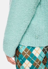 Türkis strukturierter Pullover mit lockerer Passform, der einen markierten Knopf am Saum hat. Kombiniert mit mehrfarbigen, gemusterten Karohosen.