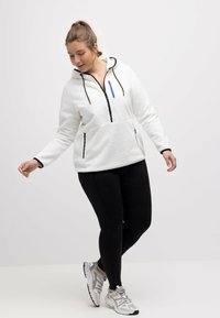 Sudadera de forro polar blanca con media cremallera, detalles en negro y cremallera azul. Combinada con leggings negros y zapatillas deportivas gris claro.