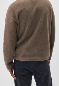 Pullover in pile marrone con una texture morbida, scollo rotondo e maniche lunghe. Abbinato a jeans scuri, che mostrano una vestibilità rilassata.