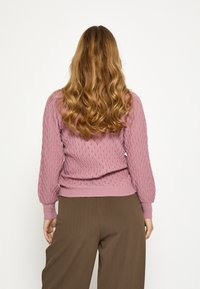 Vrouw met lang golvend blond haar, gekleed in een gestructureerde roze trui en brede bruine broek, staande tegen een effen witte achtergrond.