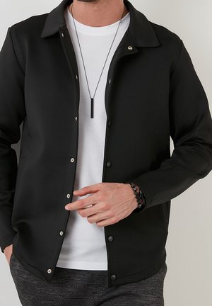 Uomo che indossa una giacca nera abbottonata sopra una camicia bianca testurizzata, collana con ciondolo nero e braccialetto nero, che con una mano sistema la giacca.