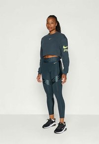 Sieviete stāv melnā īsajā garajai piedurknēm topā un melnās Nike legingās ar melniem un baltiem Nike kedām uz baltas fona.