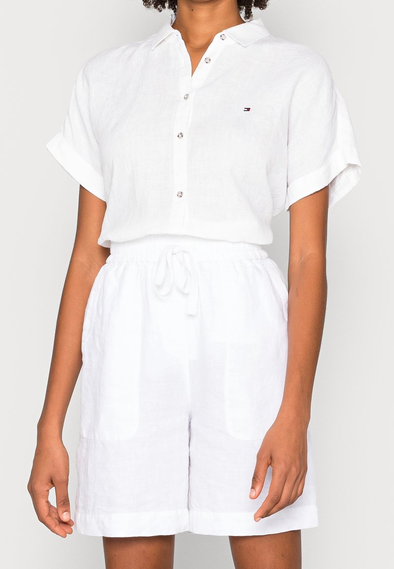 Vrouw in een witte kortmouwige overhemd met knopen en witte shorts met trekkoord voor een effen achtergrond.