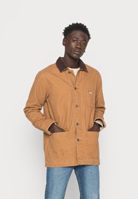 Braune Canvasjacke mit einem dunkelbraunen Cordkragen, vorderem Knopfverschluss, zwei großen Taschen und leicht lockerer Passform. Enthält sichtbare Stickdetails.