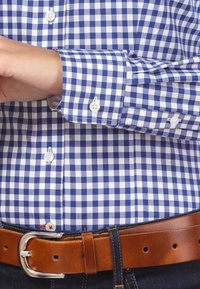 Blauw-wit geruite bloes met een knoopmanschette, gecombineerd met een bruine leren riem met een zilveren gesp, voor een achtergrond van spijkerbroeken.