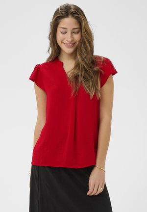 Jeune femme aux cheveux longs souriante, portant un chemisier rouge à manches courtes et une jupe noire, debout devant un fond uni.