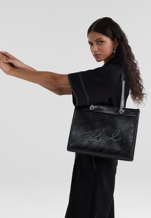 Black tote bag made of textured material with a silver chain handle, featuring studded lettering detail on the front.