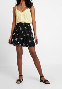 Žena oblečená v žluté bezrukávové blůze, černé květinové sukni, černých sandálech na kotníku a s vrstvami zlatých náhrdelníků, stojící na bílé podlaze.