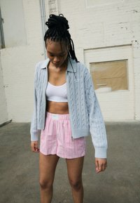 Grå kabelstickad zip-up tröja med långa ärmar över en vit magtröja, ihopparad med rosa och vita randiga shorts. Texturerade tygdetaljer synliga.