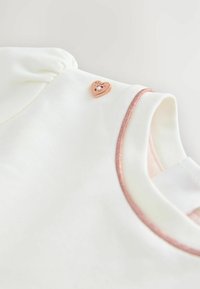 Weißes Kleidungsstück mit kurzen Puffärmel, verfügt über eine roségoldfarbene herzförmige Verzierung und einen metallisch pinken Besatz entlang des Ausschnitts.