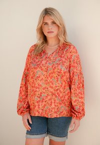 Blusa floreale di un vivace arancione, con maniche lunghe a sbuffo e vestibilità rilassata. Decorata con piccoli motivi floreali rossi e blu.