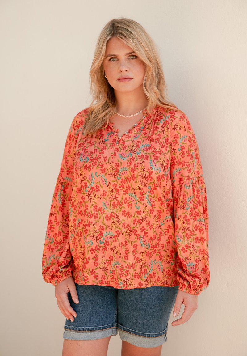 Blusa floreale di un vivace arancione, con maniche lunghe a sbuffo e vestibilità rilassata. Decorata con piccoli motivi floreali rossi e blu.