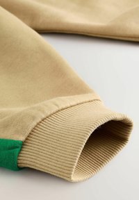 Manche de sweat-shirt beige avec bord-côte et un petit détail en tissu vert sur un fond clair.