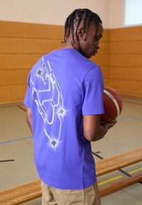 Camiseta de algodón morada con un diseño gráfico metálico plateado en la parte posterior y mangas cortas. El modelo sostiene un balón de baloncesto.