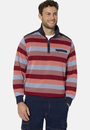 Mann trägt einen mehrfarbig gestreiften Pullover mit einem marineblauen Reißverschlusskragen und dunkelblauen Hosen, lächelt und steht mit einer Hand in der Tasche.