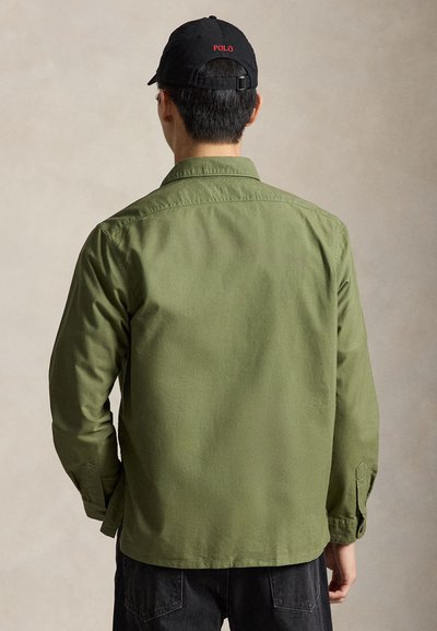 Mann trägt ein grünes Langarmshirt und eine schwarze Kappe mit dem Text "POLO", von hinten vor einem neutralen Hintergrund zu sehen.