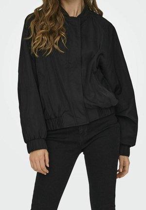 Vrouw die een zwarte bomberjack met ritssluiting draagt, voorzien van elastische manchetten en een elastische tailleband, gecombineerd met een zwarte broek, staand tegen een effen achtergrond.