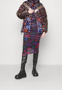 Personne portant une veste à motif multicolore, une jupe imprimée de champignons et des bottes noires à plateforme jusqu'aux genoux, se tenant devant un fond uni.