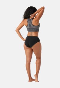 Sujetador deportivo de tela gris de punto con diseño de tirantes en la espalda, acompañado de ropa interior negra de talle alto. Modelo mostrado de espaldas, descalzo.