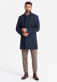 Cappotto in misto lana di colore blu marino con colletto alto, tasche laterali e dettagli con bottoni, abbinato a pantaloni a scacchi e stivali in pelle marrone.
