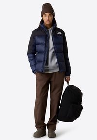 Navy and black insulated jacket, grey hoodie, brown pants, and black backpack. Slouchy brown beanie; brown, slip-on shoes.