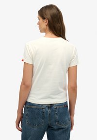 Vrouw met de rug naar de kijker, gekleed in een effen crèmekleurig T-shirt met korte mouwen en blauwe spijkerbroek, tegen een witte achtergrond.