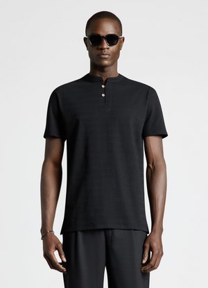 Uomo che indossa una maglia henley nera a maniche corte con due bottoni, pantaloni neri, occhiali da sole e un braccialetto al polso sinistro, in piedi su uno sfondo bianco.