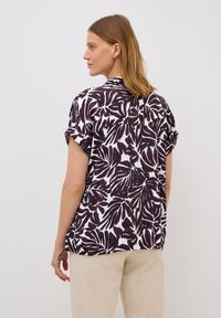 Chemise à manches courtes avec un fond blanc et des motifs de feuilles violettes foncées, coupe ample et ourlet arrondi, fabriquée en tissu léger.