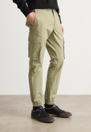 Cargo pants in light olive green, featuring an elastic waistband, tapered cuffs, and large side pockets for functionality. Styled with black sneakers.