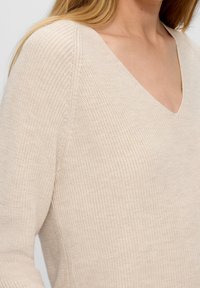 Maglione a maglia beige con ampio scollo a V e tessuto strutturato, che presenta una sottile trama a coste lungo le spalle e le maniche.