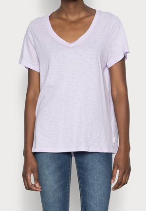 Osoba ubrana w jasnofioletowy T-shirt z dekoltem w serek i krótkim rękawem oraz niebieskie dżinsy, stojąca z rękami wzdłuż ciała.