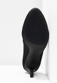 Semelle d'une chaussure à talon noir avec motif de grip texturé, posée contre un mur blanc en angle.