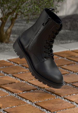 Black leather ankle boot with textured finish, round toe, lace-up design, and ridged rubber sole. Features Calvin Klein Jeans logo on side.