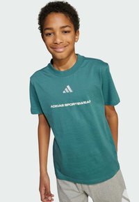 Tyrkysové bavlnené tričko s krátkymi rukávmi, okrúhlym výstrihom, s bielym textom a logom "ADIDAS SPORTSWEAR". Skombinované so sivými šortkami.